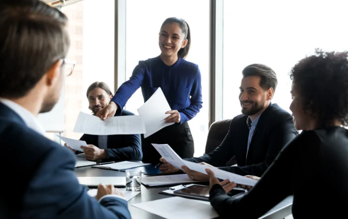 Female team leader share handouts at office meeting | Quick Reliable Printing (QRP) A woman in a blue blouse hands out papers to colleagues at a round table, smiling as they review documents in a bright office.Quick Reliable Printing (QRP)