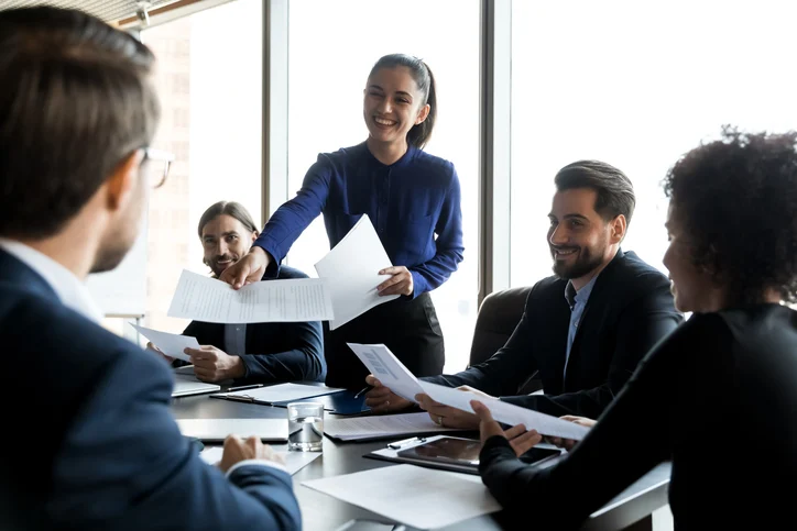 A woman in a blue blouse hands out papers to colleagues at a round table, smiling as they review documents in a bright office.Quick Reliable Printing (QRP)