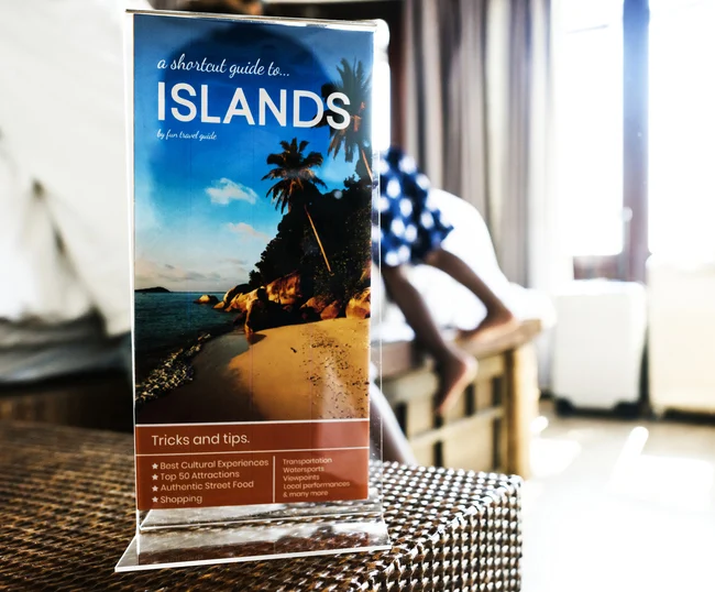 Travel brochure standing upright on a woven table, showing a beach scene with palm trees and the large title 'ISLANDS'.Quick Reliable Printing (QRP)