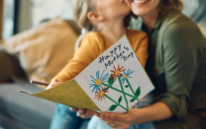 Close-up of woman receiving Mothers day greeting card from her daughter | Quick Reliable Printing (QRP) A child and mother kiss while the child holds a handmade Mother's Day card with colorful flowers on display.Quick Reliable Printing (QRP)