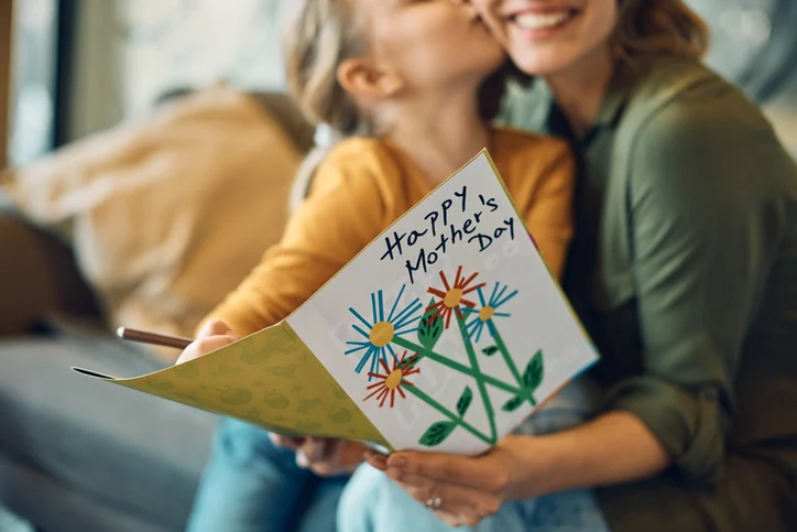 A child and mother kiss while the child holds a handmade Mother's Day card with colorful flowers on display.Quick Reliable Printing (QRP)