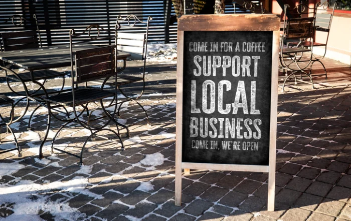 Support Local Business Advertising Sign Outside Cafe On Winter Street | Quick Reliable Printing (QRP) Chalkboard sign outside a cafe reads 'Support Local Business' with outdoor metal tables and snow on the cobblestone ground.Quick Reliable Printing (QRP)
