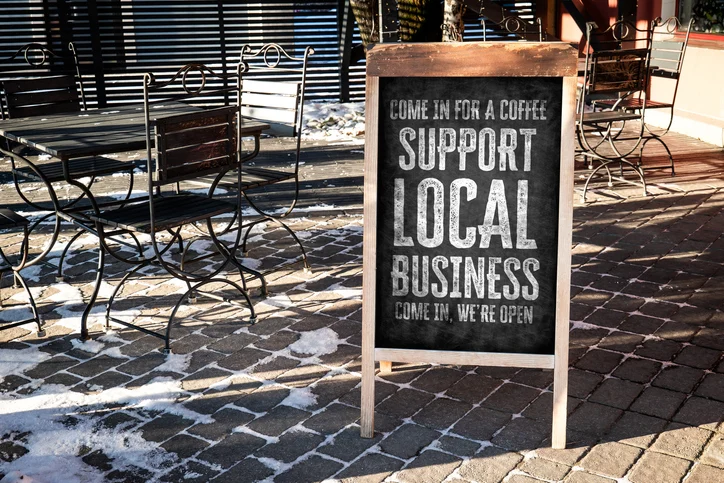 Chalkboard sign outside a cafe reads 'Support Local Business' with outdoor metal tables and snow on the cobblestone ground.Quick Reliable Printing (QRP)
